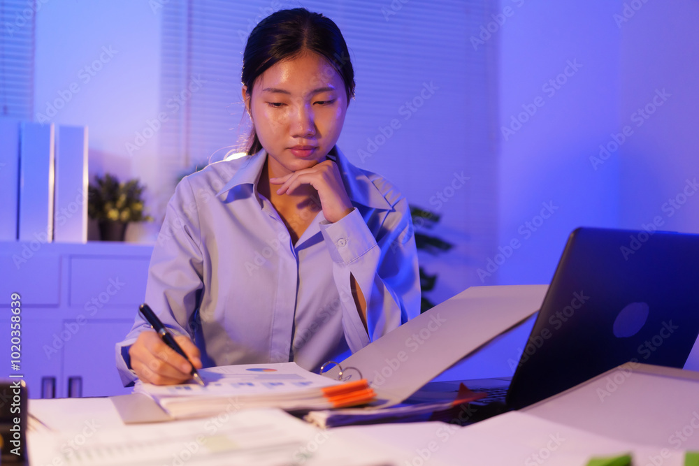 An Asian woman is working overtime at her desk in the office, busy viewing documents and her notebook. She is talking while managing tasks late at night, staying focused and productive An Asian woman is working overtime at her desk in the office, busy viewing documents and her notebook. She is talking while managing tasks late at night, staying focused and productive