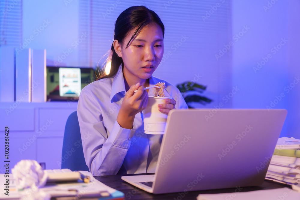 An Asian woman works late at her desk in the office, eating instant noodles while looking at documents and working on her laptop. She is busy with overtime submissions and business tasks at night