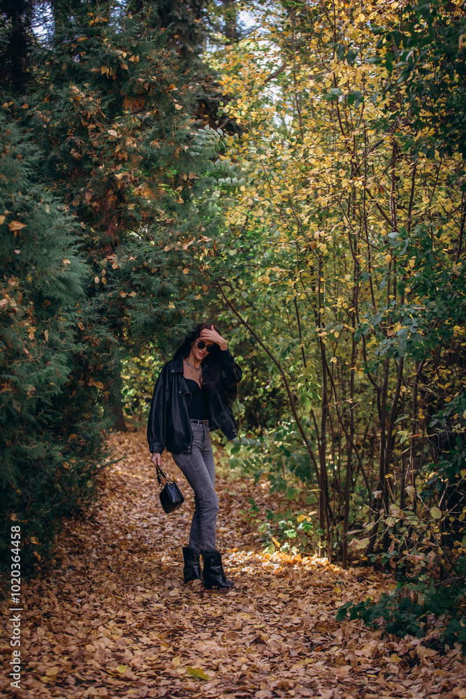 Woman, an adult and a beautiful brunette in a leather jacket and jeans, walks in the park in autumn. Woman, an adult and a beautiful brunette in a leather jacket and jeans, walks in the park in autumn.