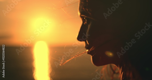 Close up woman face against bright orange sunset. Girl silhouette enjoys amazing colorful sunset, standing on a tropical beach, raises hands against orange dramatic sky and ocean waves. Holiday travel