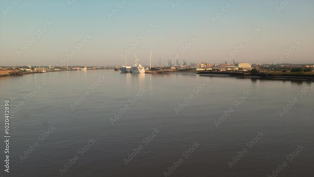 Slow-motion aerial shot of the Thames River at sunrise, showcasing Tilbury Docks with cruise ships, wind turbines, and industrial port in the background near London. Slow-motion aerial shot of the Thames River at sunrise, showcasing Tilbury Docks with cruise ships, wind turbines, and industrial port in the background near London.