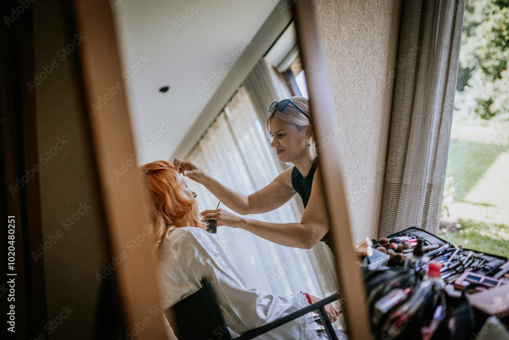 A makeup artist applies makeup to a woman with red hair, captured in a relaxed and natural setting with window light streaming in. A makeup artist applies makeup to a woman with red hair, captured in a relaxed and natural setting with window light streaming in.