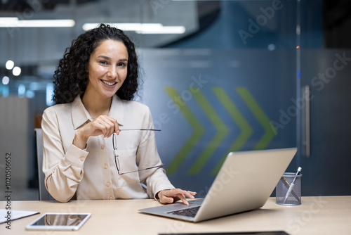 Smiling woman with glasses sitting at desk using laptop. Concept of professionalism, confidence, business environment. Office supplies, phone, and laptop present. Bright corporate setting.