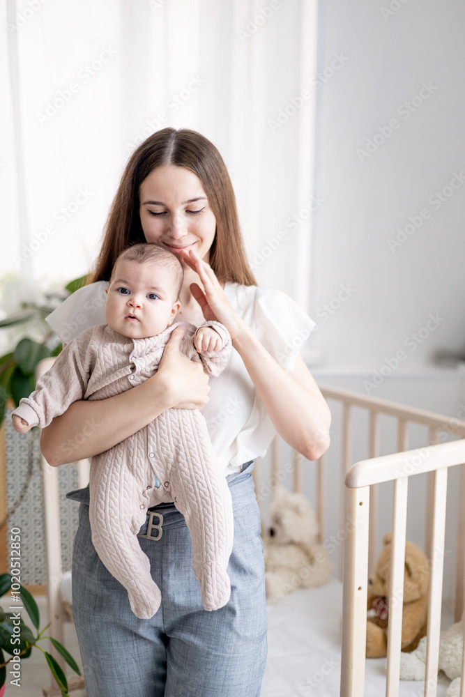 a young mother gently embraces her little baby girl holding him in her arms at home in the bedroom of the nursery by the bed, close-up. Portrait of a happy mother and a newborn baby. a young mother gently embraces her little baby girl holding him in her arms at home in the bedroom of the nursery by the bed, close-up. Portrait of a happy mother and a newborn baby.