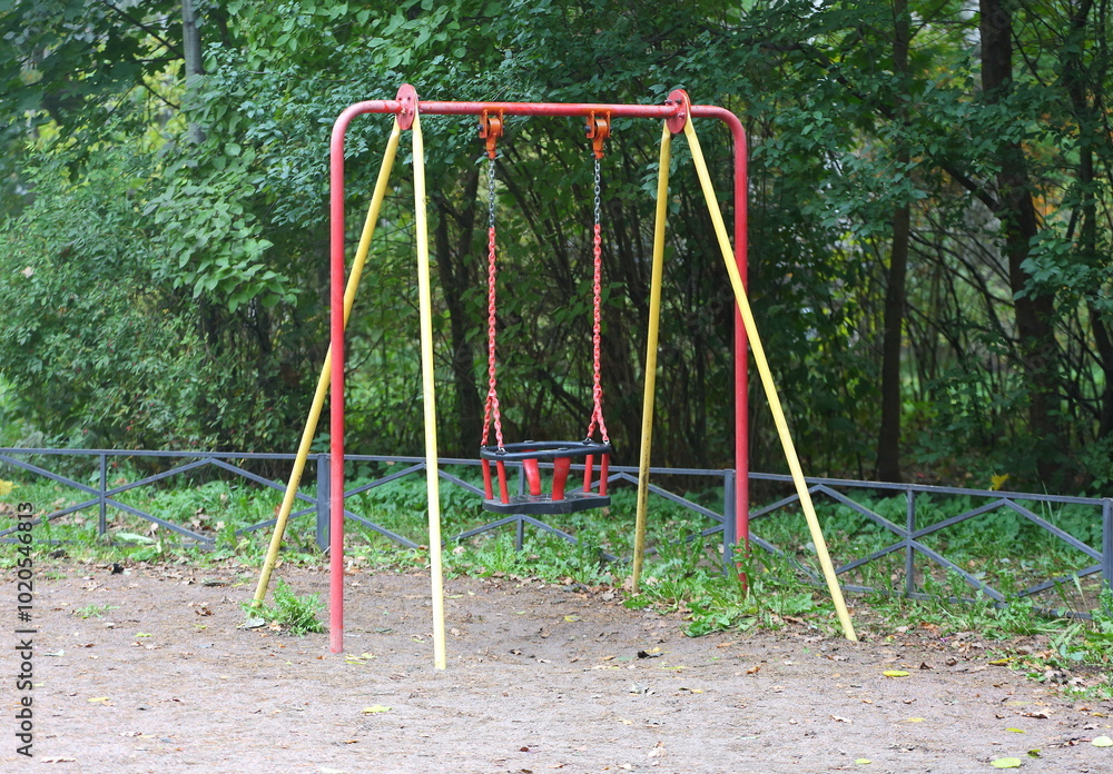 A children's swing stands near green bushes surrounded by a low fence A children's swing stands near green bushes surrounded by a low fence