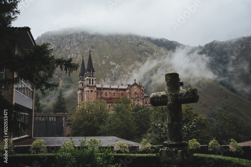 Stone cross is standing in a garden with the basilica of covadonga in the background, surrounded by fog and mountains photo