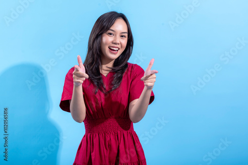 A young Asian woman wearing a red dress stands confidently against a light blue background, smiling and pointing with both hands. The playful gesture and vibrant colors create a cheerful