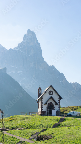 Alpine church on a green field with a rugged mountain peak in the background on a sunny day in Passo Rolle, Dolomites