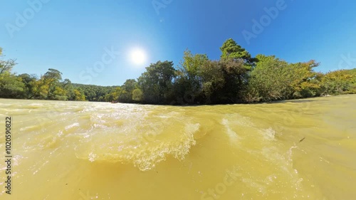 A scenic view of a muddy river under a bright blue sky, with lush green trees lining the shore, captured from a low angle on the water's surface, emphasizing the sun's reflection and natural surroundi photo