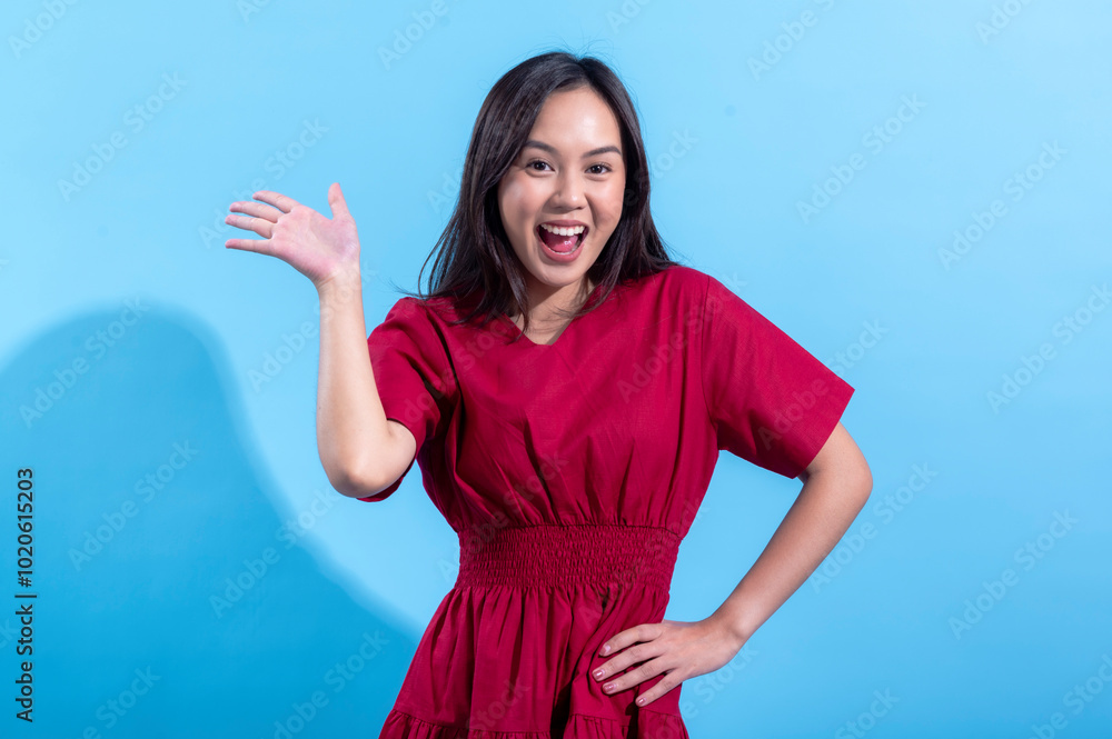 An Asian woman in a red dress waves cheerfully with her right hand raised while smiling widely. She stands confidently with one hand on her hip, posing against a light blue background An Asian woman in a red dress waves cheerfully with her right hand raised while smiling widely. She stands confidently with one hand on her hip, posing against a light blue background