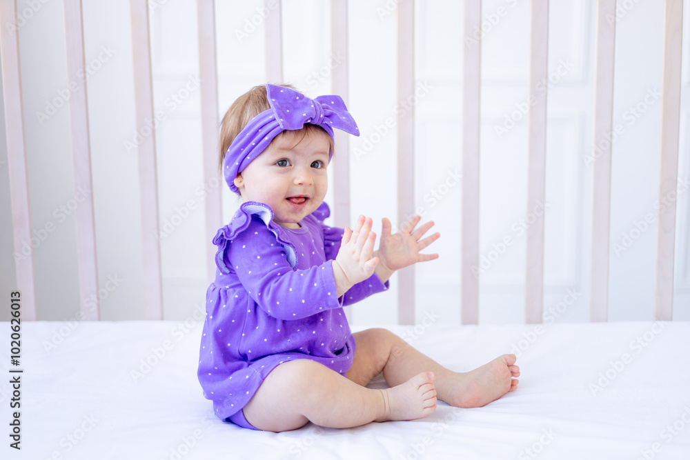 cute baby girl sitting on the bed in lilac clothes and with a bow on her head and smiling, funny little baby on a cotton bed at home playing, the concept of children's goods cute baby girl sitting on the bed in lilac clothes and with a bow on her head and smiling, funny little baby on a cotton bed at home playing, the concept of children's goods