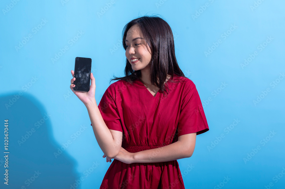 Asian woman in a red dress holds a smartphone in her outstretched hand, presenting it to the camera with a smile. against a light blue background, showcasing the phone screen Asian woman in a red dress holds a smartphone in her outstretched hand, presenting it to the camera with a smile. against a light blue background, showcasing the phone screen