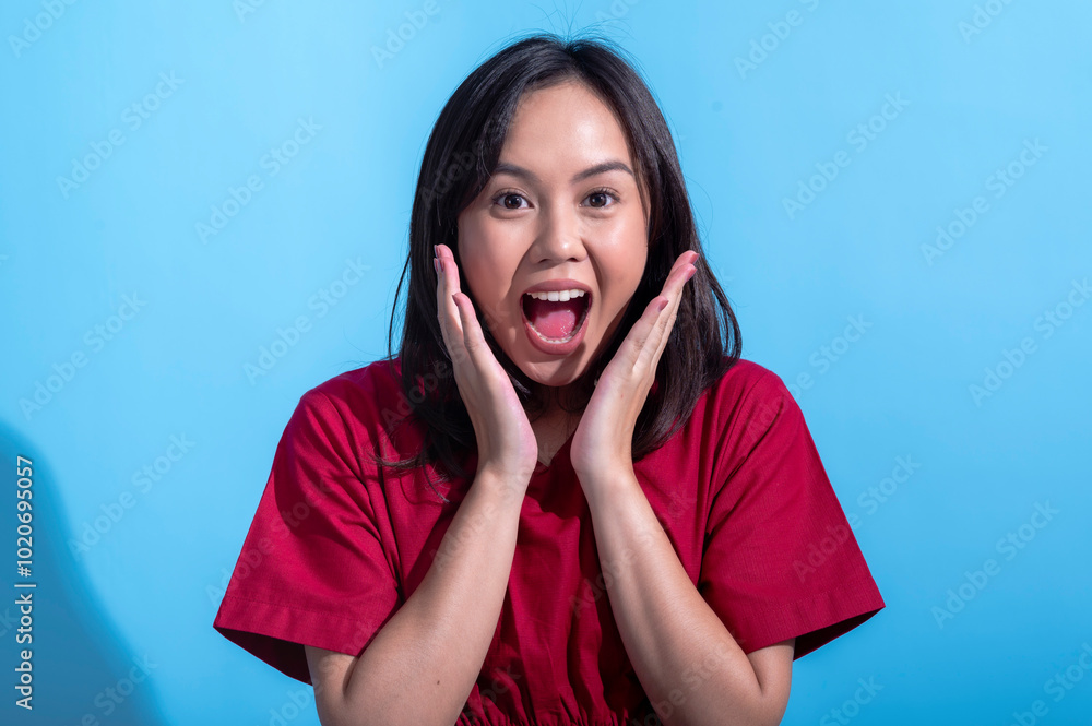 An Asian woman in a red dress looks shocked with wide eyes and both hands pressed against her cheeks. She expresses surprise and disbelief. The background is light blue An Asian woman in a red dress looks shocked with wide eyes and both hands pressed against her cheeks. She expresses surprise and disbelief. The background is light blue
