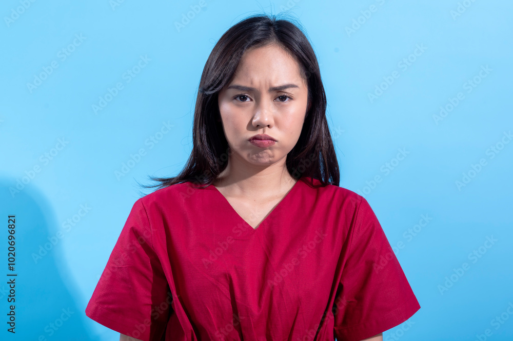 An Asian woman wearing a red dress is expressing frustration or displeasure, with her lips puffed out and brows furrowed. She stands against a light blue background, which contrasts with her outfit An Asian woman wearing a red dress is expressing frustration or displeasure, with her lips puffed out and brows furrowed. She stands against a light blue background, which contrasts with her outfit