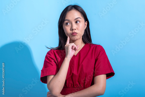 Asian woman in a red dress stands against a light blue background, looking thoughtful with her finger on her cheek. She appears to be pondering, with a curious expression on her face