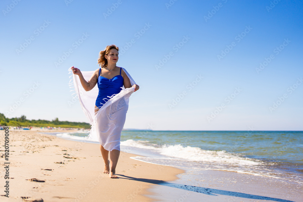 Summer vacations. Beautiful young woman in blue swimsuit covered with white scarf walking on sand by sea on beach on sunny day. Summer vacations. Beautiful young woman in blue swimsuit covered with white scarf walking on sand by sea on beach on sunny day.