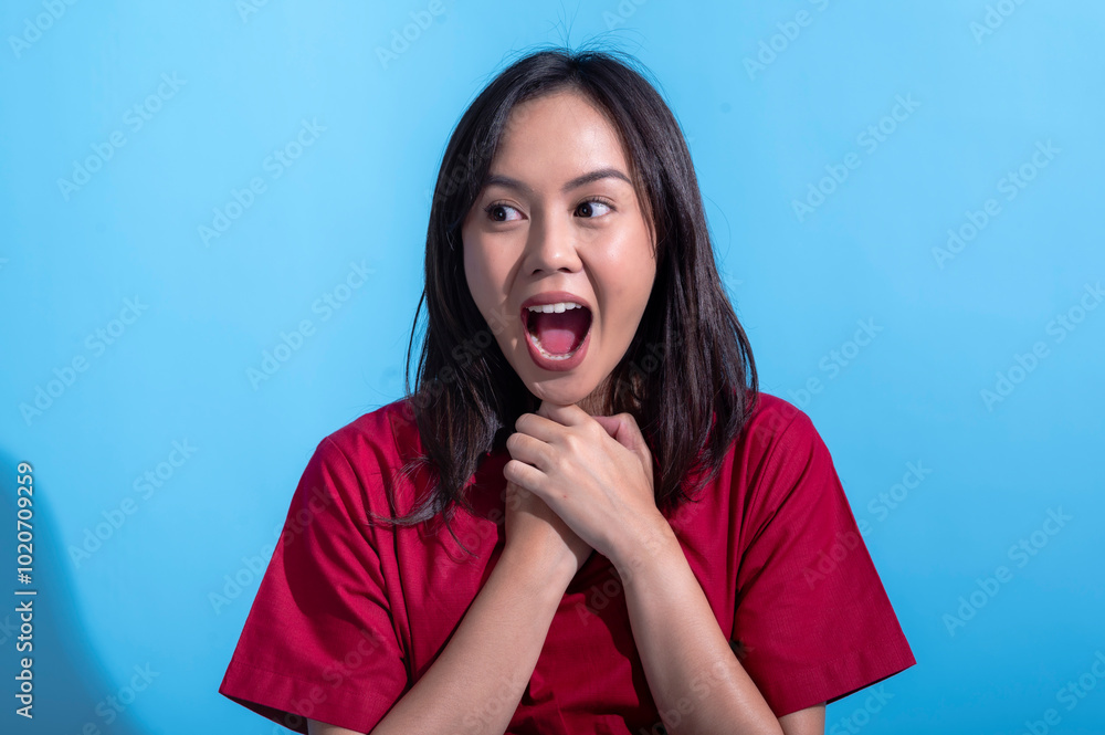 An Asian woman in a red dress stands against a light blue background with her mouth wide open, expressing shock or surprise. Her hands are clasped near her neck, emphasizing her astonishment An Asian woman in a red dress stands against a light blue background with her mouth wide open, expressing shock or surprise. Her hands are clasped near her neck, emphasizing her astonishment