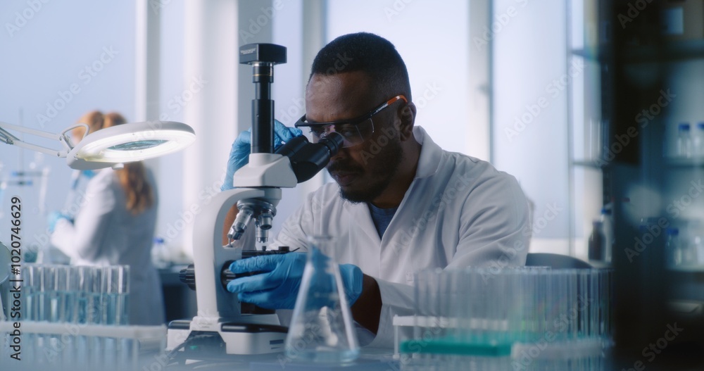 Scientists in white coats work in advanced laboratory. African American microbiologist examines liquid from test tubes with samples under microscope. In the background, female scientist does analysis. Scientists in white coats work in advanced laboratory. African American microbiologist examines liquid from test tubes with samples under microscope. In the background, female scientist does analysis.