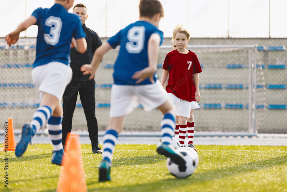 Fototapeta premium Group of boys attending soccer football training on a school grass pitch. Man coaching children in physical education class. Soccer practice for teenage boys. Soccer players in red and blue uniforms