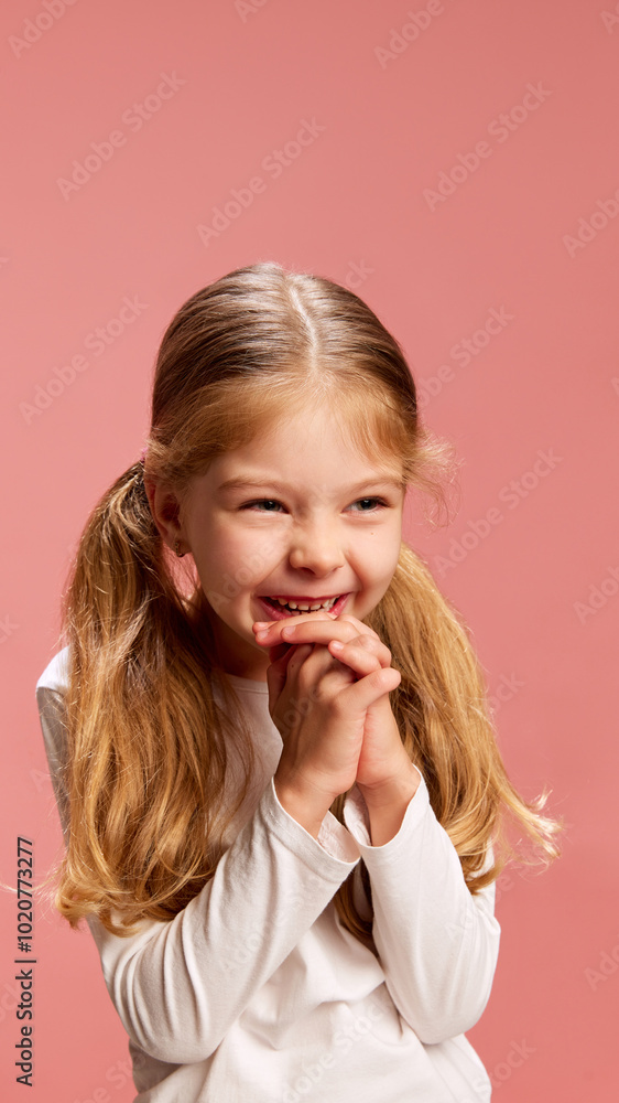 Little girl smiles slyly with her hands clasped under her chin, giving off sweet and bashful vibe against pastel pink background. Concept of innocence, joy, human emotions, childhood. Ad