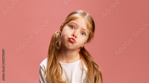Cute little girl creates pouty, playful expression, lips puckered and her head tilted, giving off sense of silly defiance against pastel pink background. Concept of human emotions, childhood, positive