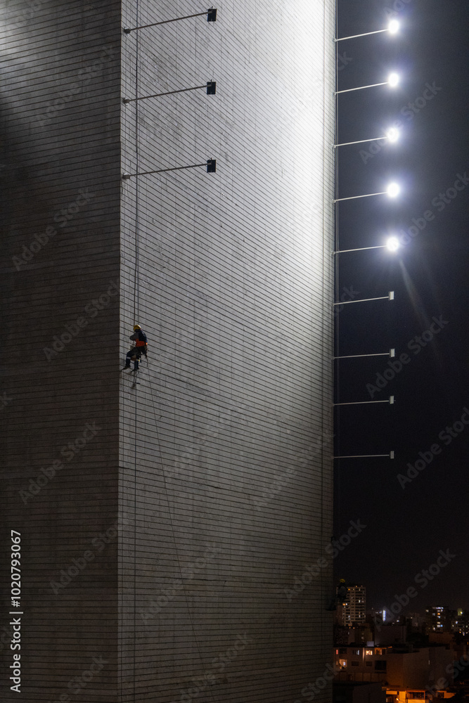 Man Hanging Up Working on the Wall of a Building Under Construction at Night with Horizontal Lights on Poles, Vertical Picture