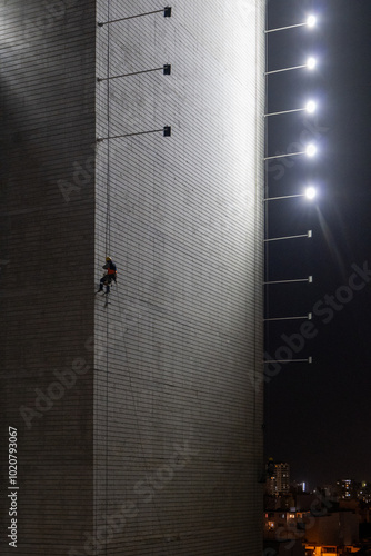Obraz na płótnie Man Hanging Up Working on the Wall of a Building Under Construction at Night with Horizontal Lights on Poles, Vertical Picture w salonie