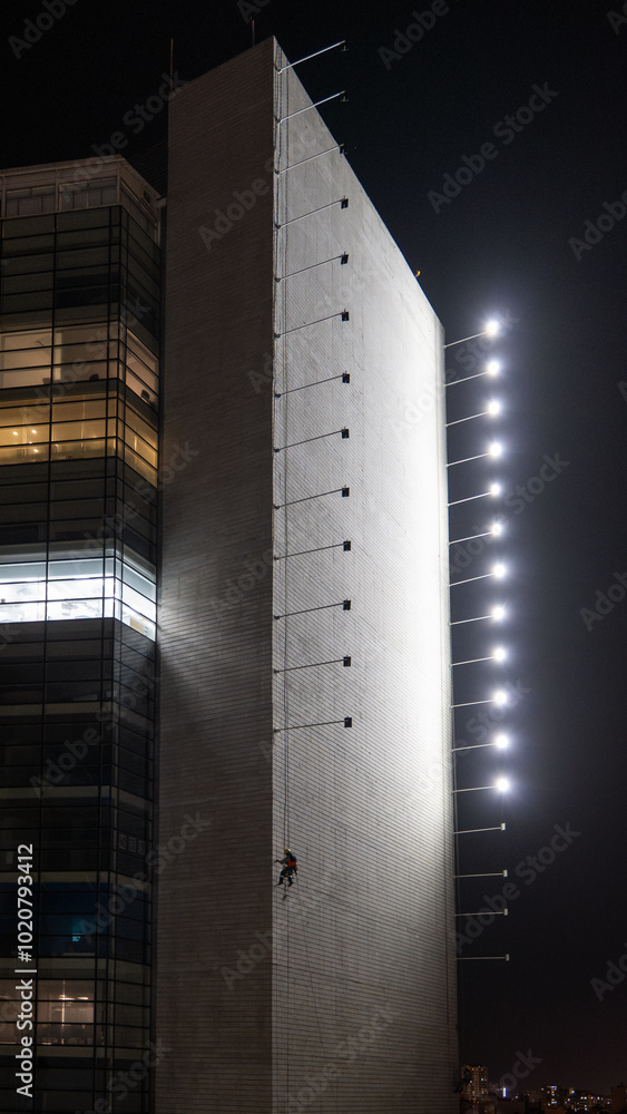 Man Hanging Up Working on the Wall of a Building Under Construction at Night with Horizontal Lights on Poles, Vertical Picture