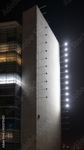 Obraz na płótnie Man Hanging Up Working on the Wall of a Building Under Construction at Night with Horizontal Lights on Poles, Vertical Picture w salonie