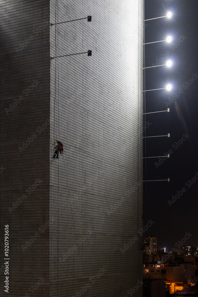 Man Hanging Up Working on the Wall of a Building Under Construction at Night with Horizontal Lights on Poles, Vertical Picture