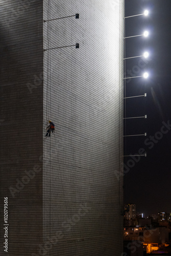 Obraz na płótnie Man Hanging Up Working on the Wall of a Building Under Construction at Night with Horizontal Lights on Poles, Vertical Picture w salonie