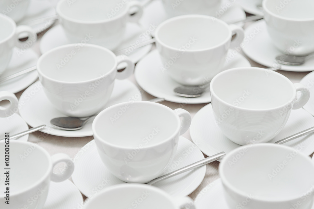 white empty porcelain cups on saucers with spoons in several rows on white tablecloth, serving tables for banquet in restaurant or cafe, pattern, background white empty porcelain cups on saucers with spoons in several rows on white tablecloth, serving tables for banquet in restaurant or cafe, pattern, background