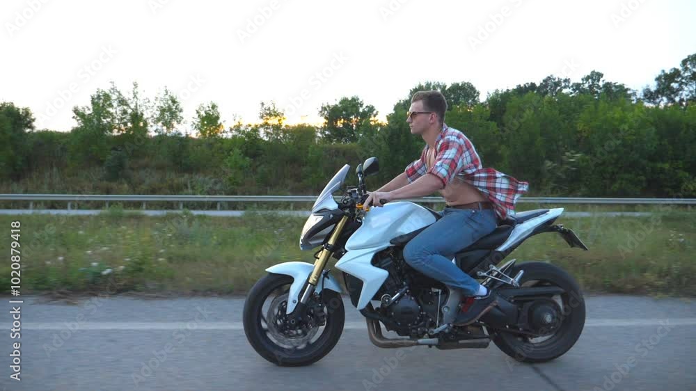 Young man riding on a modern sport motorbike. Handsome motorcyclist in a shirt and glasses driving his motorcycle on country road with sunflowers field at background. Slow motion Side view Close up Young man riding on a modern sport motorbike. Handsome motorcyclist in a shirt and glasses driving his motorcycle on country road with sunflowers field at background. Slow motion Side view Close up