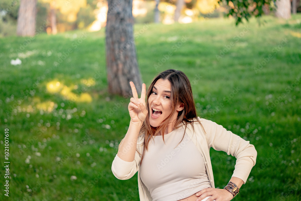 Beautiful young woman is making the peace sign with her fingers while smiling in a park Beautiful young woman is making the peace sign with her fingers while smiling in a park