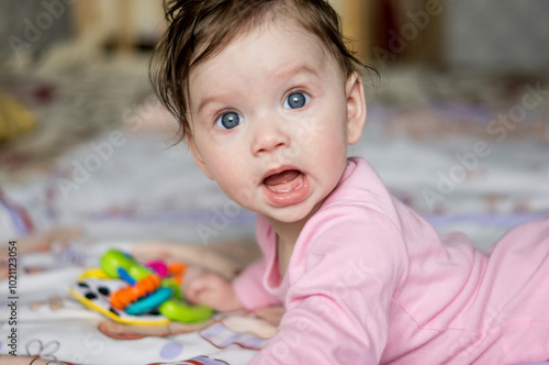 A small child or baby or toddler or girl lies on her stomach, looks at the camera with surprise or fear, opening her toothless mouth. Portrait of a child with big blue eyes