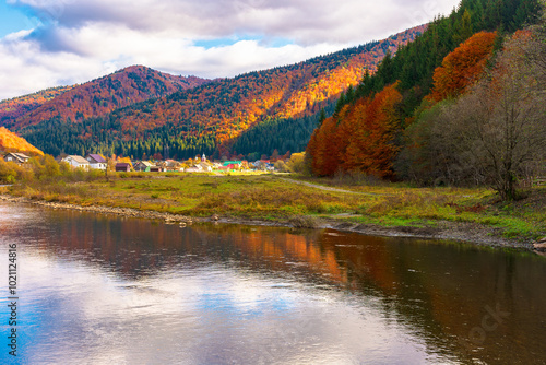 autumn landscape with tereblya river in carpatian mountains. countryside scenery of ukraine on a cloudy day in fall season reflecting in the calm water. village in the distance. colorful woods