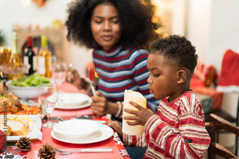 Playful African boy blowing decorated candle at dining table during Christmas festive dinner with family. people enjoy at xmas party together. Playful African boy blowing decorated candle at dining table during Christmas festive dinner with family. people enjoy at xmas party together.