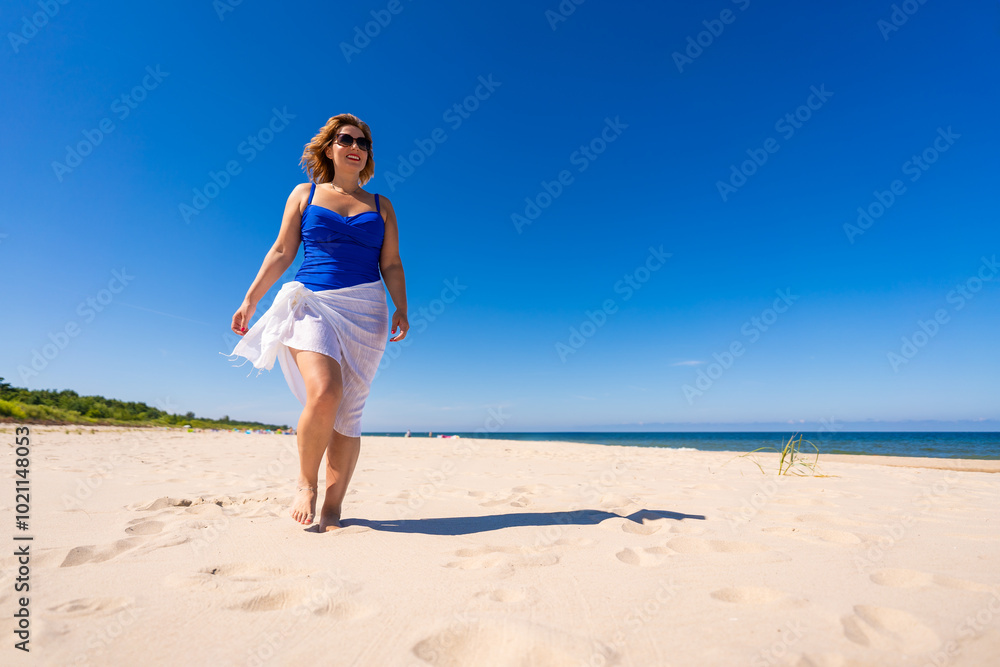 Summer vacation. Beautiful young woman in one-piece blue swimsuit and white scarf and sunglasses walking on sandy beach on beautiful sunny day. Sunbathing. Summer vacation. Beautiful young woman in one-piece blue swimsuit and white scarf and sunglasses walking on sandy beach on beautiful sunny day. Sunbathing.