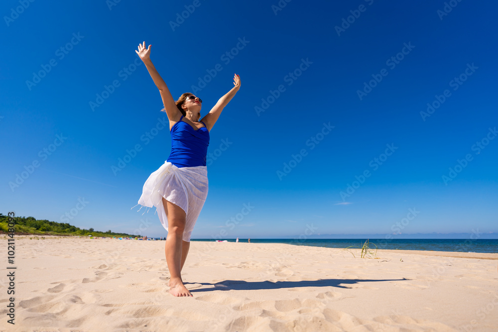 Summer vacation. Beautiful young woman in one-piece blue swimsuit and white scarf and sunglasses standing with her arms raised up on sandy beach on beautiful sunny day. Freedom and power. Summer vacation. Beautiful young woman in one-piece blue swimsuit and white scarf and sunglasses standing with her arms raised up on sandy beach on beautiful sunny day. Freedom and power.