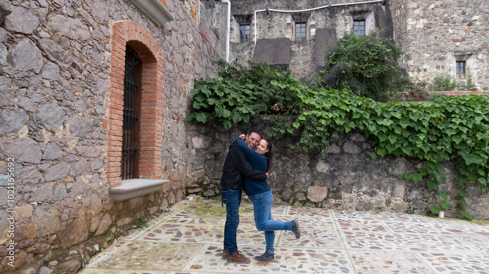 pareja hombre y mujer se dan abrazo y mian a la camara sonrientes con paisaje rural al fondo, ventana de ladrillo, muro de piedra y follaje pareja hombre y mujer se dan abrazo y mian a la camara sonrientes con paisaje rural al fondo, ventana de ladrillo, muro de piedra y follaje