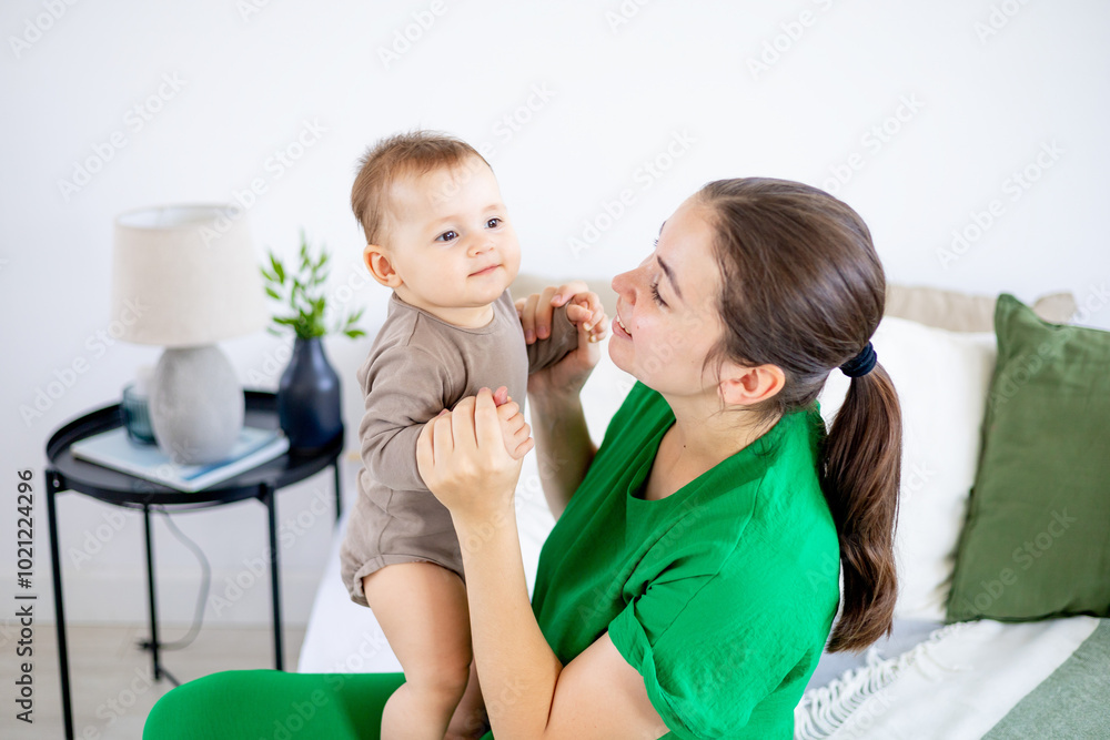 a young mother in green clothes gently hugs a baby boy on a bed in a bright bedroom, happy motherhood a young mother in green clothes gently hugs a baby boy on a bed in a bright bedroom, happy motherhood