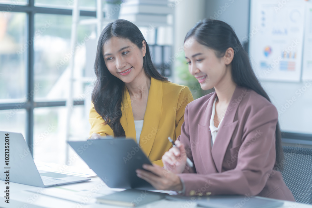 Two young businesswomen smiling, reviewing a contract in a modern office. Working together, focused on discussion, showcasing professionalism. Bright space with laptop and documents Two young businesswomen smiling, reviewing a contract in a modern office. Working together, focused on discussion, showcasing professionalism. Bright space with laptop and documents
