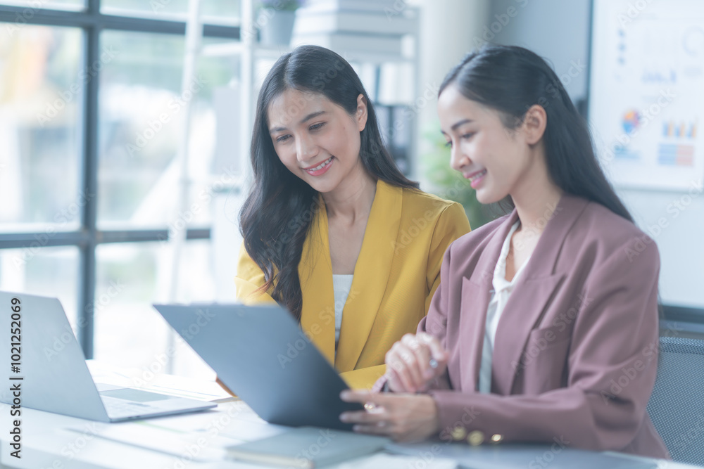 Two young businesswomen collaborating and discussing documents in a stylish office, using laptops to analyze data and charts for their startup Two young businesswomen collaborating and discussing documents in a stylish office, using laptops to analyze data and charts for their startup