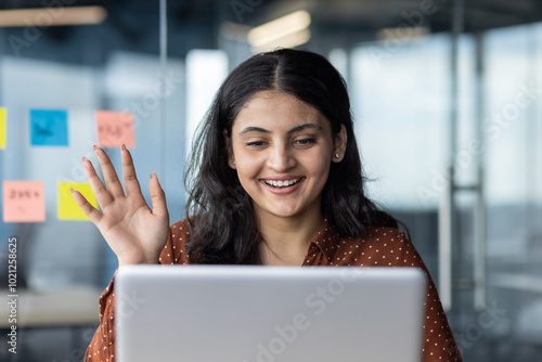 Smiling woman waves at laptop screen during virtual meeting, showcasing positive communication in modern office. Sticky notes on glass wall complement her cheerful engagement in digital workspace.