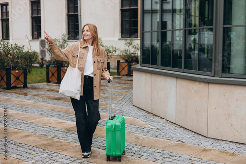 Cheerful young woman in casual outfit walking on the city street with green luggage. 30s Woman with suitcase pointing finger on urban background. Travel Fashion. Full body