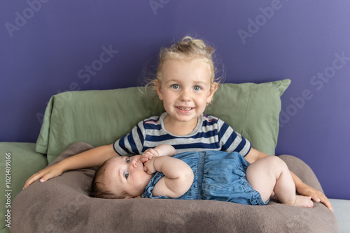 Smiling girl with long blond hair lovingly holds her baby sister on her lap, creating a heartwarming scene of family bonding and togetherness. Selective focus photo