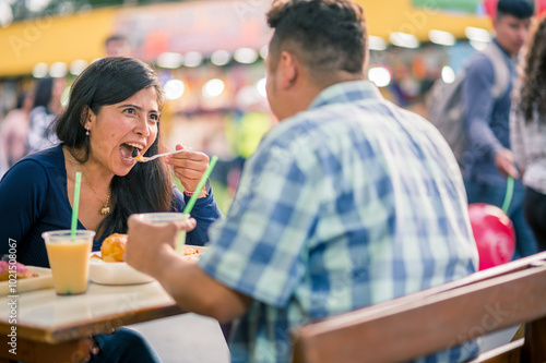 Latina woman brings a bite of food to her mouth, she is sitting in front of her boyfriend at a street food place.