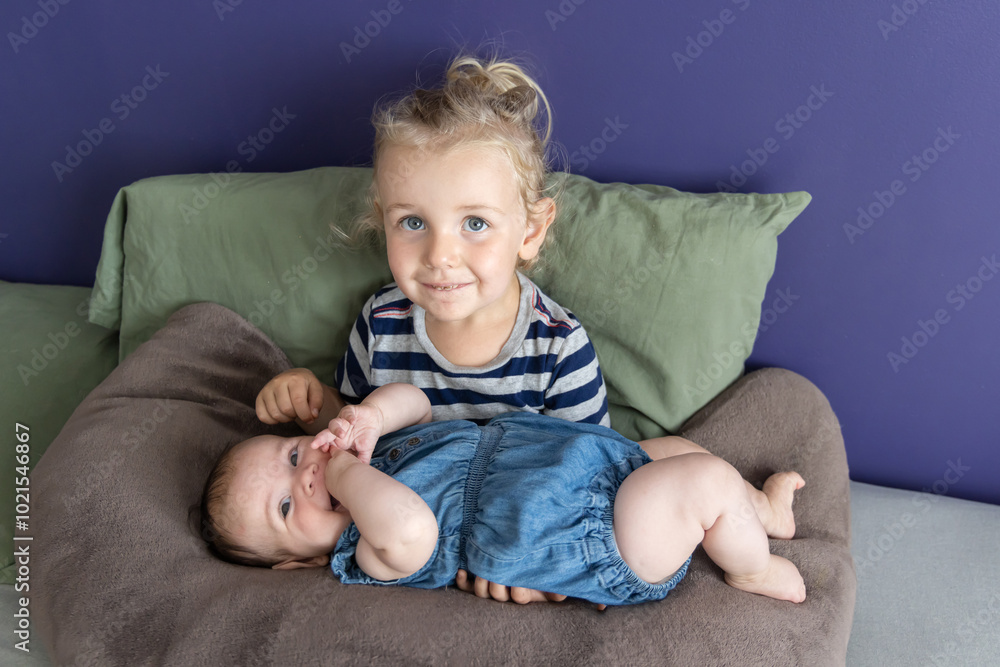 Young child is holding their baby sister on their lap while sitting on a bed with a purple wall. Selective focus Young child is holding their baby sister on their lap while sitting on a bed with a purple wall. Selective focus