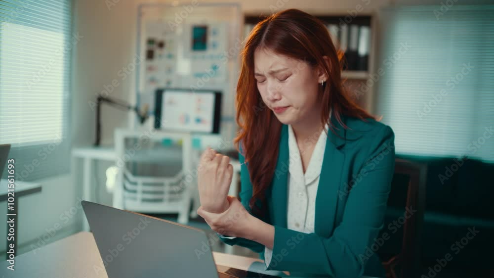 Asian businesswoman in a green suit massages her wrist while working at her desk, showing signs of pain and discomfort from prolonged computer use and stress in the office environment Asian businesswoman in a green suit massages her wrist while working at her desk, showing signs of pain and discomfort from prolonged computer use and stress in the office environment