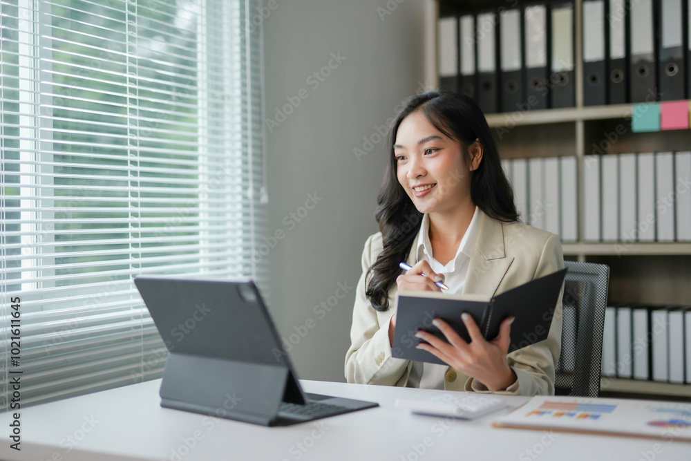Businesswoman is taking notes in a notebook while having a work video call on a tablet in her office Businesswoman is taking notes in a notebook while having a work video call on a tablet in her office